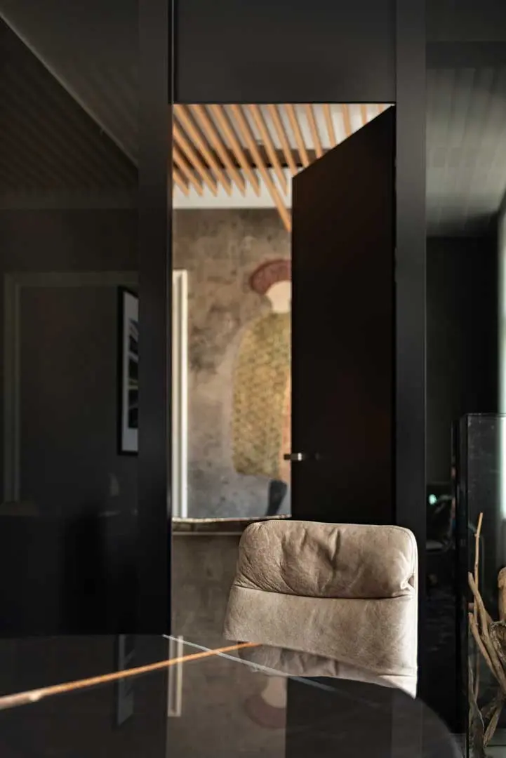 Ein beiger Stuhl steht vor einem glänzenden schwarzen Tisch in einem modernen Raum mit dunklen Wänden. Eine offene schwarze Tür gibt den Blick auf eine Wandmalerei frei, die eine Figur mit Hut und gemustertem Mantel zeigt. Ein Beispiel gelungener Office-Fotografie.