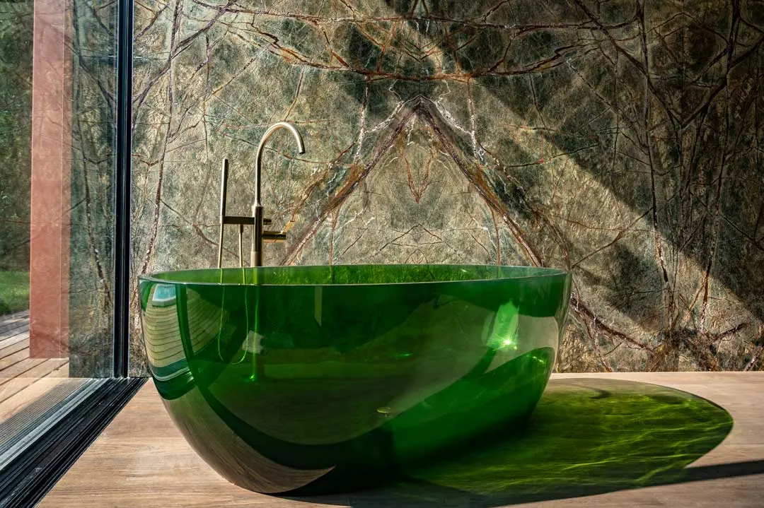 Ein modernes Badezimmer mit einer durchscheinenden grünen Badewanne, einem eleganten goldenen Wasserhahn und einer großen Marmorwand mit verschlungenen natürlichen Mustern. Durch eine Glastür strömt Sonnenlicht herein und wirft grüne Reflexe auf den Boden.