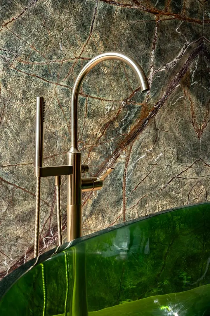 Ein moderner Wasserhahn in gebürstetem Gold und eine Handbrause sind vor einer grün-braun marmorierten Wand neben einer glänzenden, grün durchscheinenden Badewanne montiert.