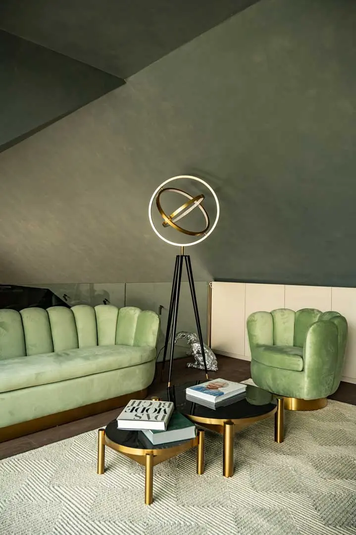 Ein modernes Wohnzimmer mit einem grünen Samtsofa und -sessel, runden schwarz-goldenen Couchtischen mit Büchern, einer geometrischen Stehlampe und einem hellen, strukturierten Teppich darunter. Die Wände und die Decke sind in gedeckten Tönen gestrichen.