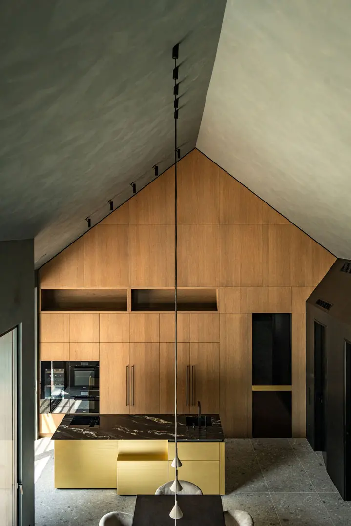 Moderne Interiorfotografie einer Küche mit hoher Gewölbedecke, Holzschränken und minimalistischer Pendelbeleuchtung. In der Mitte befindet sich eine dunkle Kücheninsel mit integriertem Kochfeld und gelbem Sockel, im Vordergrund steht ein Esstisch.