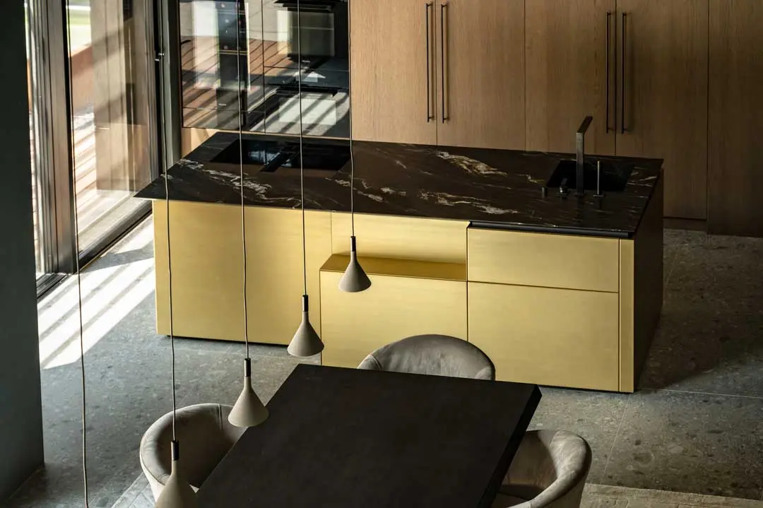 Moderne Küche in einer Villa mit einer Insel aus schwarzem Marmor, goldenen Schränken, Einbaugeräten, Holzschränken und Pendelleuchten über einem dunklen Esstisch mit beigen Stühlen. Sonnenlicht strömt durch große Fenster.