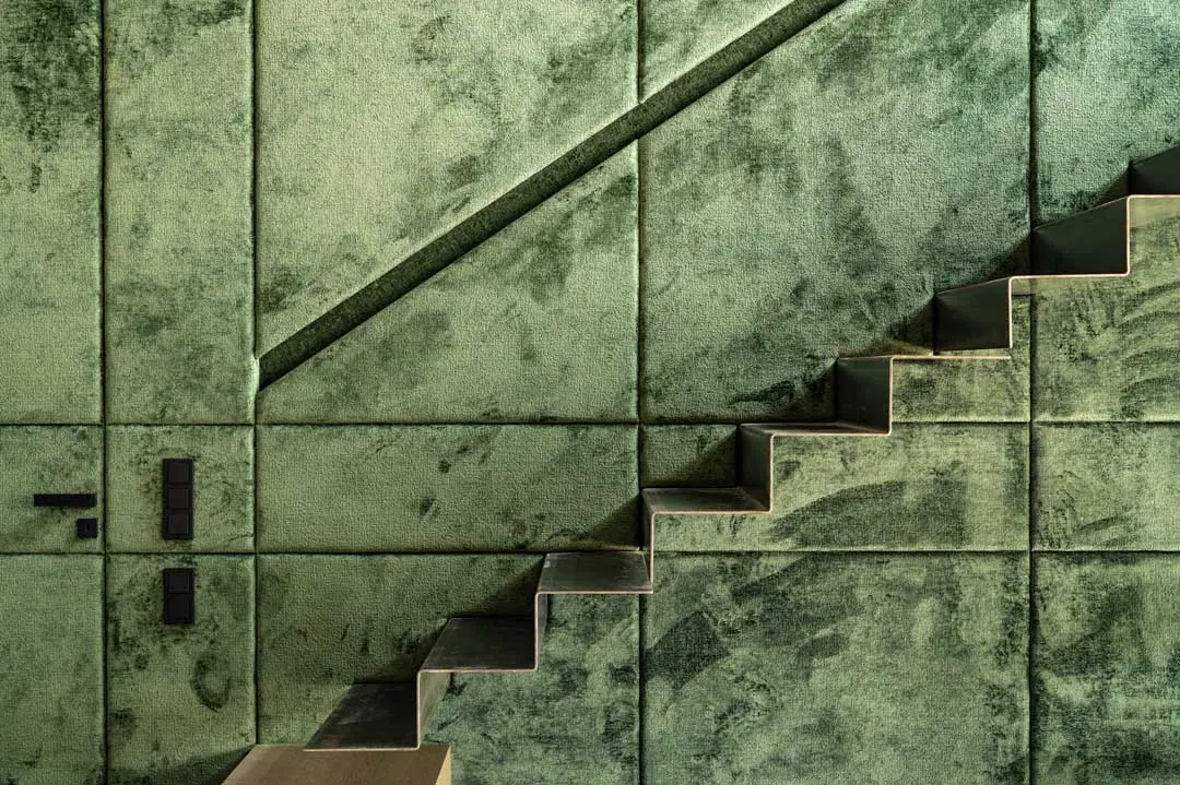 Eine moderne Treppe mit scharfen, kantigen Stufen führt vor einer grünen, strukturierten Wand mit großen, samtigen Paneelen und geometrischen Linien nach oben und schafft so ein markantes architektonisches Design. Eine professionelle Interiorfotografie für ein Architekturbüro.