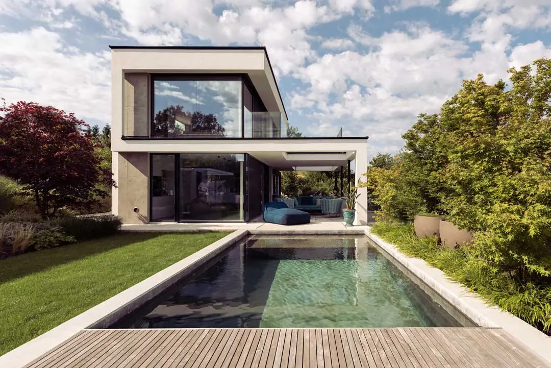Modernes zweistöckiges Tiny House mit großen Glasfenstern, einem üppig begrünten Garten, einer Holzterrasse und einem rechteckigen Swimmingpool davor, umgeben von Bäumen und Pflanzen unter einem teilweise bewölkten Himmel.