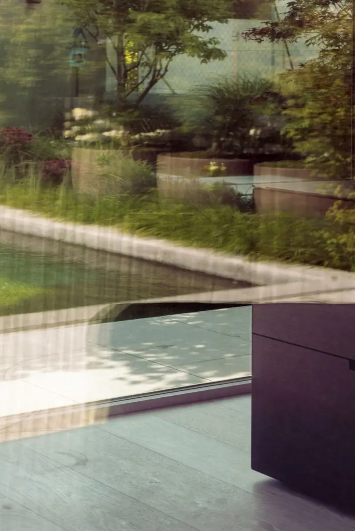 In einem modernen Glasfenster spiegelt sich ein üppiger Garten mit grünen Pflanzen und Bäumen sowie ein rechteckiger Swimmingpool und eine Steinterrasse. Das Interieur hat klare Linien und minimalistische dunkle Möbel. Ein echtes Wunschhaus.